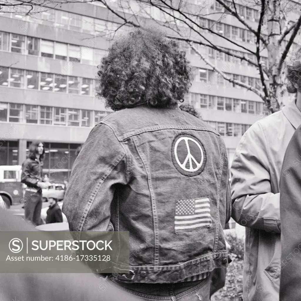 1960s 1970s ANONYMOUS MALE HIPPIE BACK VIEW LONG CURLY HAIR BLUE JEAN DENIM JACKET AMERICAN FLAG AND PEACE SYMBOL PATCHES