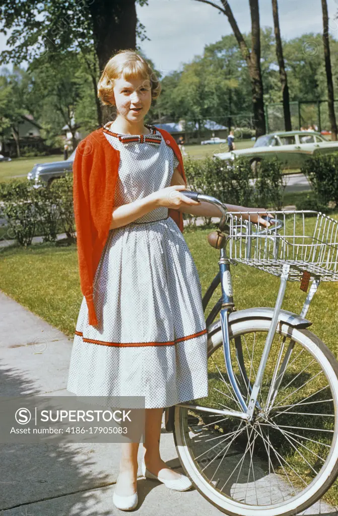 1950s BLOND TEENAGE GIRL WEARING DRESS AND RED CARDIGAN SWEATER STANDING WITH BICYCLE LOOKING AT CAMERA