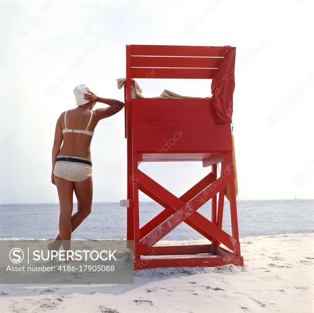 1980s REAR VIEW ANONYMOUS WOMAN IN WHITE BIKINI LEANING AGAINST LIFEGUARD STATION BEACH CHAIR BARNEGAT NEW JERSEY USA