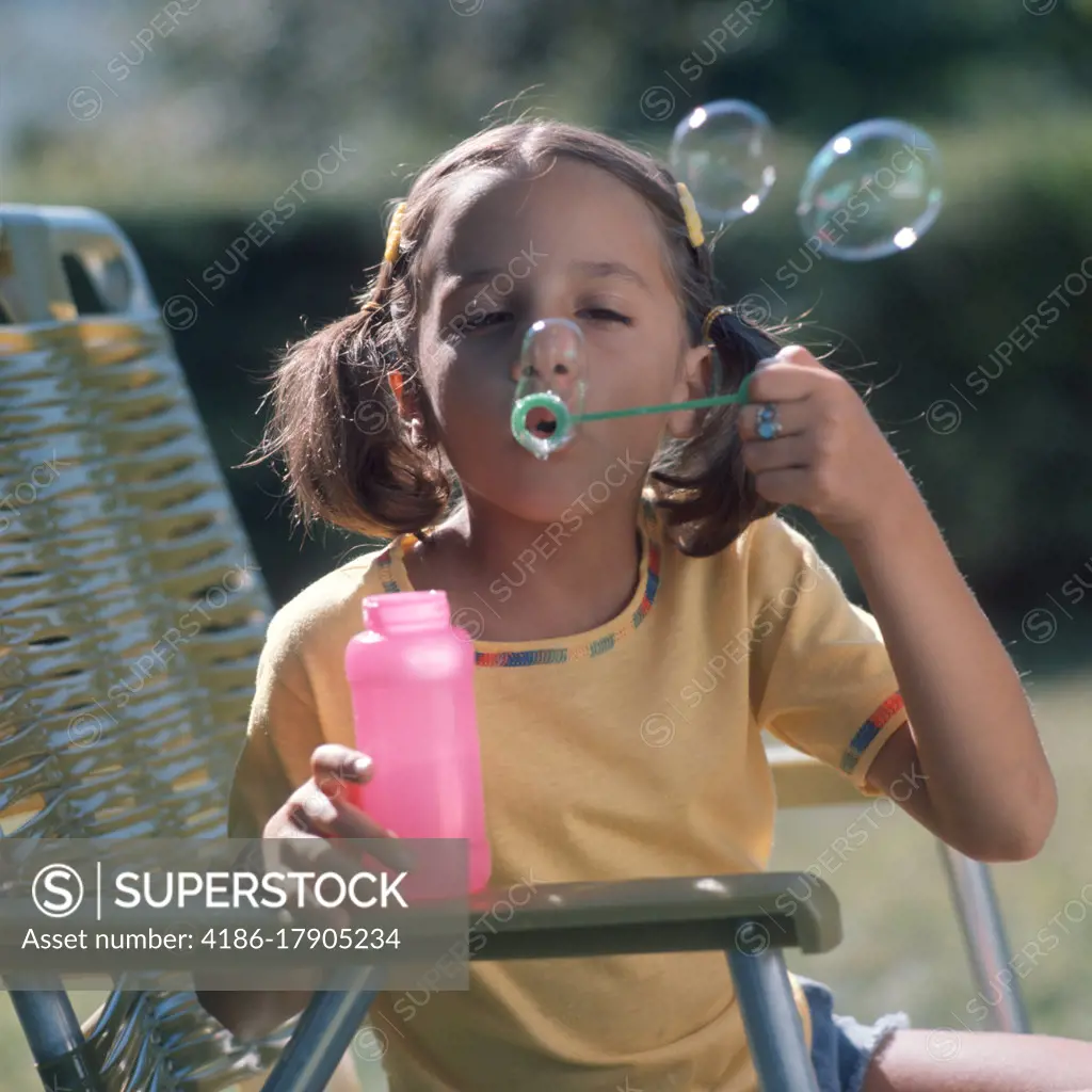 1970s GIRL IN PIGTAILS YELLOW SHIRT SITTING ON LAWN CHAIR BLOWING SOAP BUBBLES FROM SOAPY WATER SOLUTION IN PINK PLASTIC BOTTLE