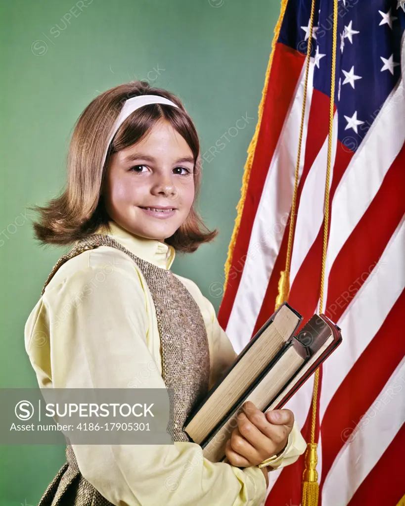 1960s SMILING GIRL WEARING TWEED JUMPER YELLOW BLOUSE HOLDING TEXTBOOKS SMILING LOOKING AT CAMERA STANDING BESIDE AMERICAN FLAG