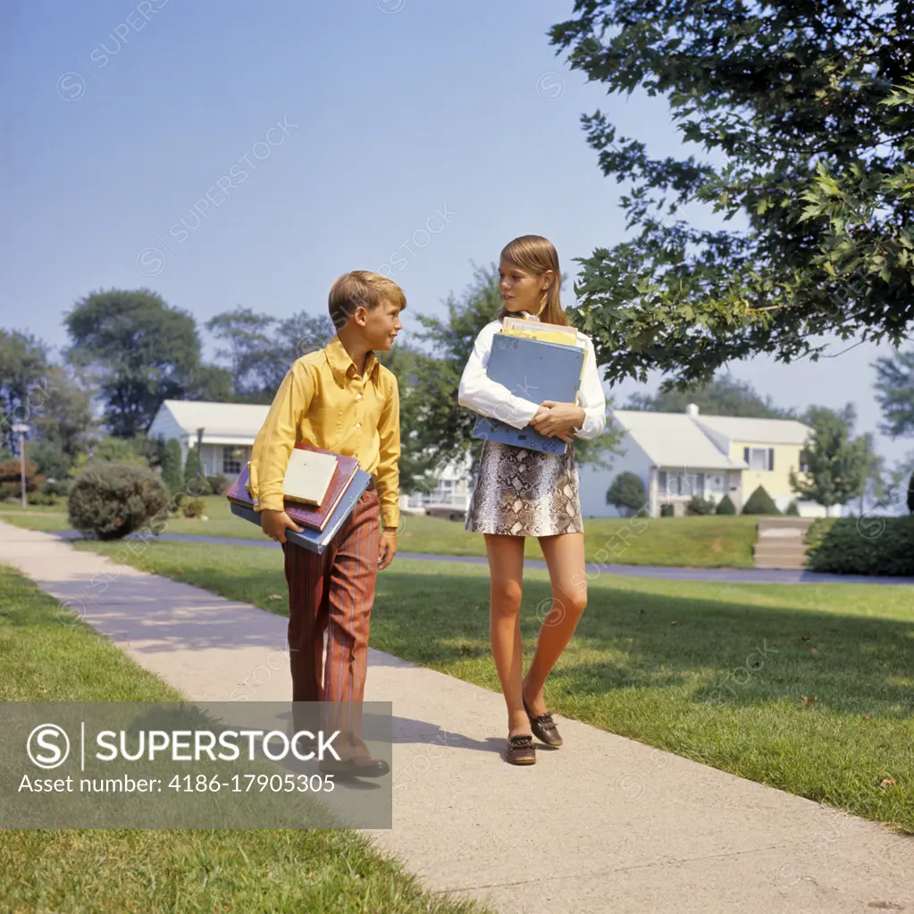 1960s 1970s BOY GIRL ON SIDEWALK WALKING HOME FROM OR TO SCHOOL TALKING CARRYING BOOKS GIRL WEARING FAUX SNAKESKIN MINI SKIRT