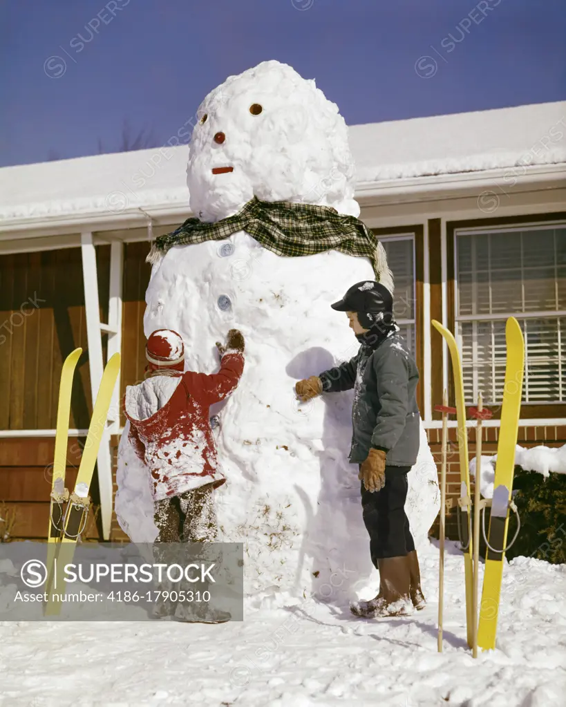 1960s TWO BOYS WITH VERY TALL SNOWMAN BY HOUSE YELLOW SKIS STUCK IN SNOW