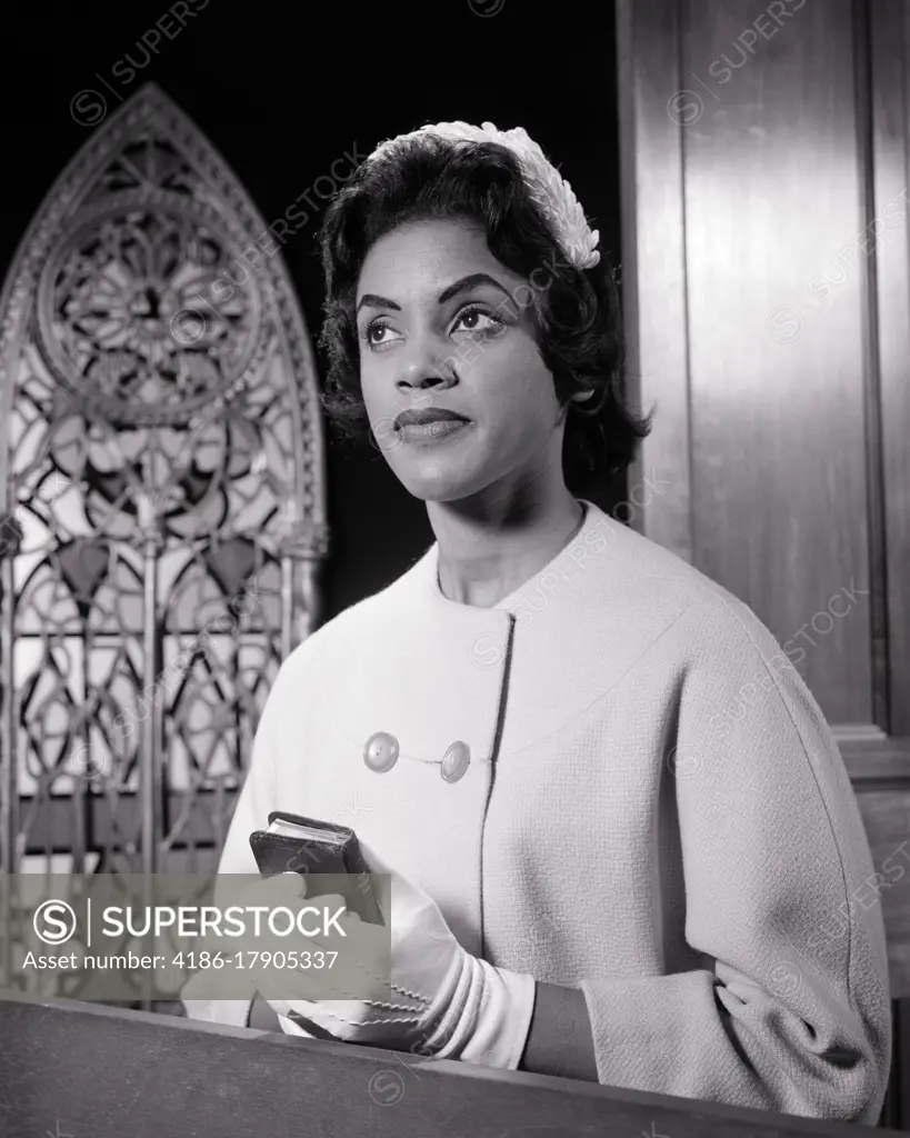 1960s AFRICAN-AMERICAN WOMAN IN CHURCH PEW WEARING WHITE HAT AND GLOVES HOLDING A SMALL BIBLE PRAYER BOOK