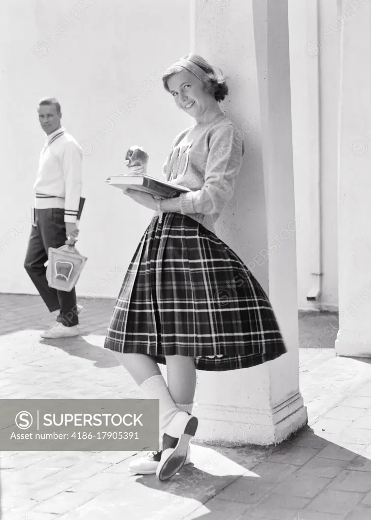 1950s 1960s TEEN GIRL LEANING AGAINST COLUMN EATING APPLE READING BOOK LOOKING AT CAMERA SMILING AS POTENTIAL BOYFRIEND WALKS BY