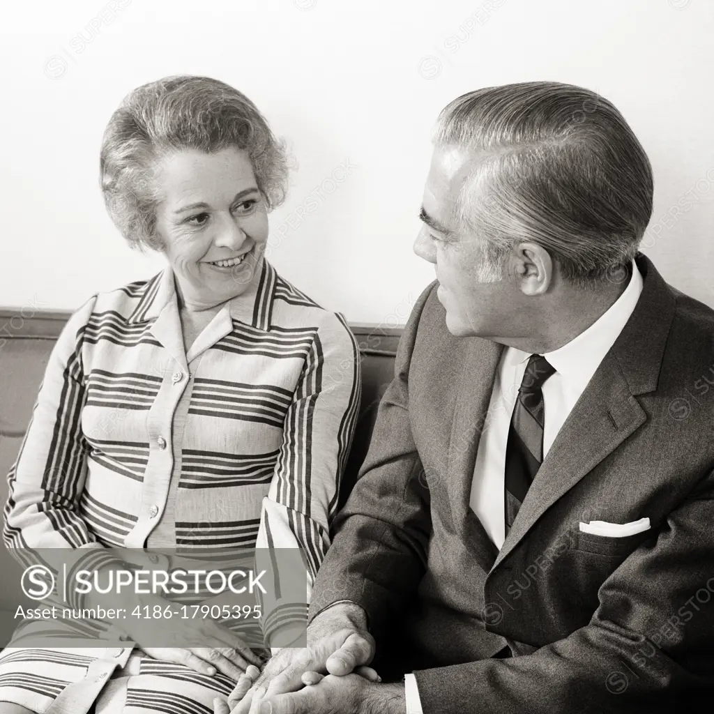 1960s 1970s SENIOR MATURE COUPLE SITTING ON COUCH HOLDING HANDS LOOKING AT EACH OTHER