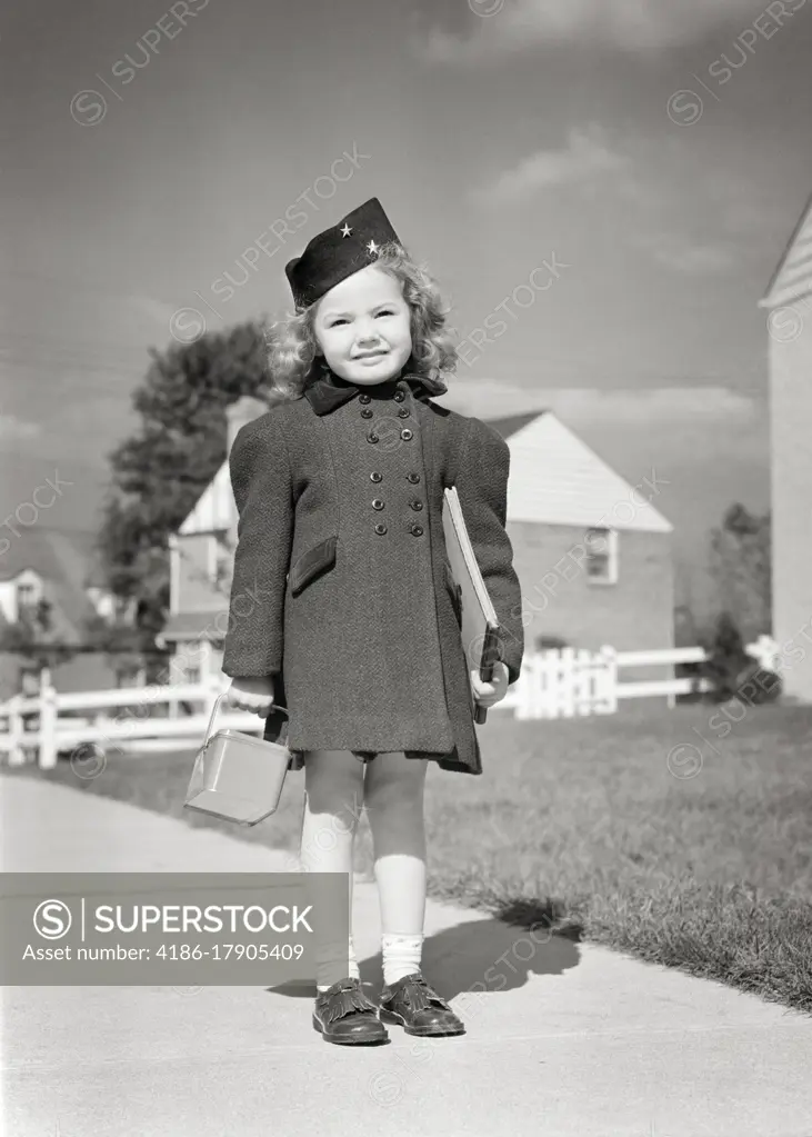 1940s SMILING LITTLE GIRL READY FOR SCHOOL STANDING ON SIDEWALK WEARING BLACK HAT DOUBLE BREASTED COAT HOLDING LUNCH BOX & BOOK