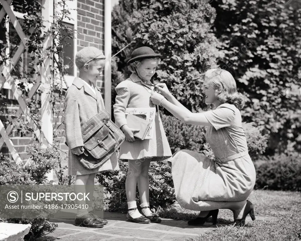 1940s 1950s MOTHER ADJUSTING CHECKING COAT ON GIRL AND BOY DAUGHTER SON SISTER BROTHER READY TO LEAVE FOR SCHOOL BOOK BAG BOOKS