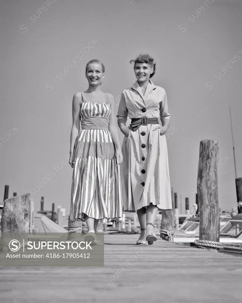 1940s 1950s TWO SMILING YOUNG WOMEN WEARING STYLISH SUMMER DRESSES WALKING SIDE-BY-SIDE ON WOODEN BOAT DOCK AT A MARINA 