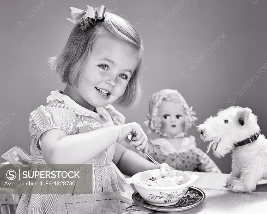 1940s 1950s SMILING GIRL BOW IN HAIR EATING BOWL OF ICE CREAM LOOKING AT CAMERA SURROUNDED BY STUFFED ANIMAL DOG AND DOLL TOYS 