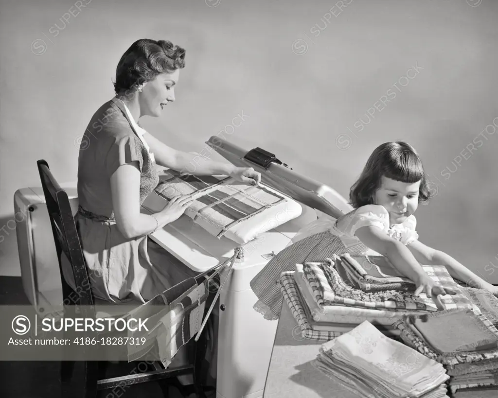 1950s WOMAN HOUSEWIFE USING AN ELECTRIC APPLIANCE STEAM PRESS TO IRON LINENS AS GIRL DAUGHTER FOLDS AND STACKS THEM ON TABLE