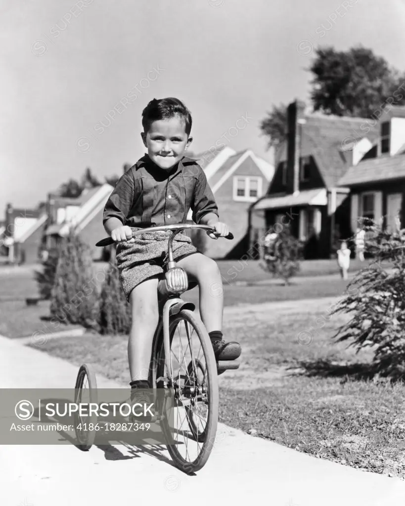 1930s 1940s BOY RIDING TRICYCLE BIKE ON SUBURBAN SIDEWALK SMILING HAPPY DETERMINED FACIAL EXPRESSION LOOKING AT CAMERA 