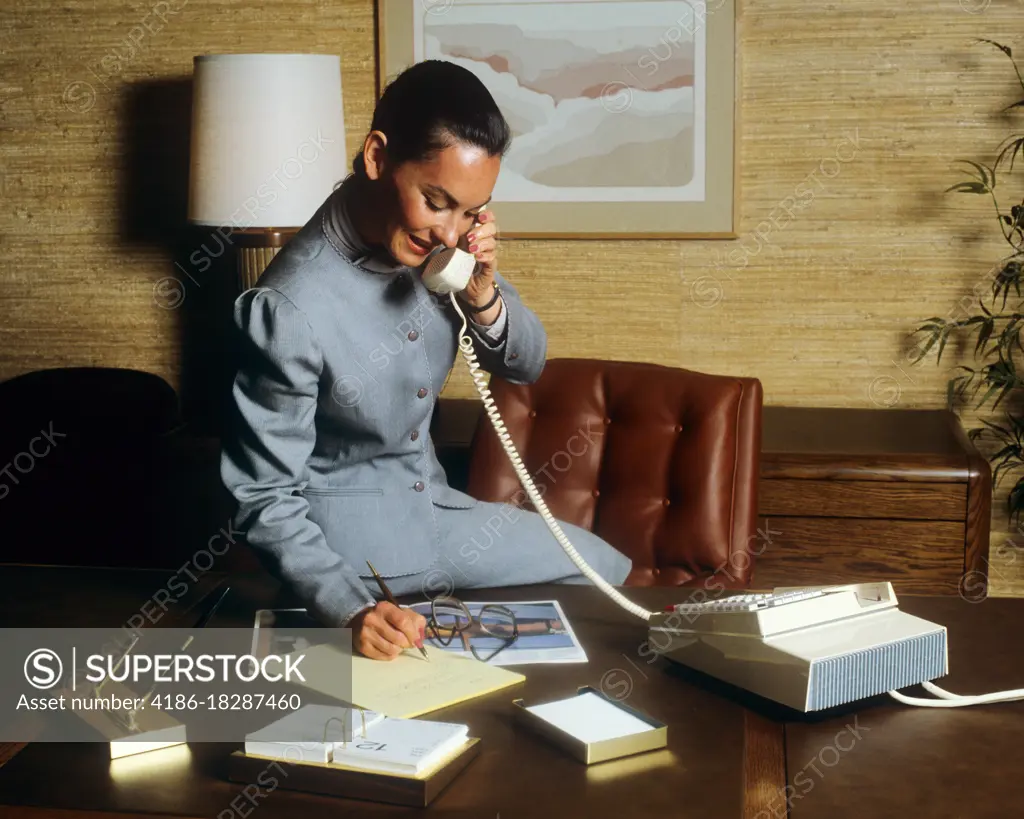 1980s SMILING BUSINESS WOMAN IN OFFICE SITTING ON HER DESK TALKING ON TELEPHONE WRITING NOTES