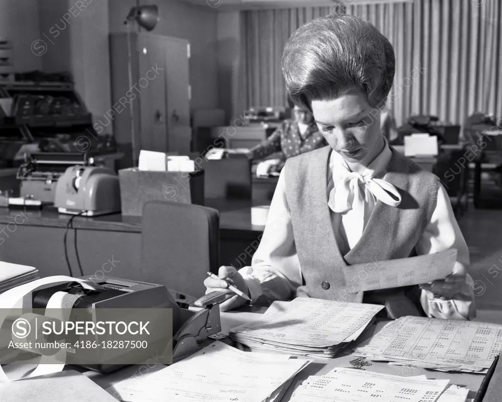 1960s OFFICE WORKER WOMAN WITH TEASED BIG HAIR HAIRSTYLE WORKING AT DESK WITH ADDING MACHINE AND HUGE PILES OF PAPERS