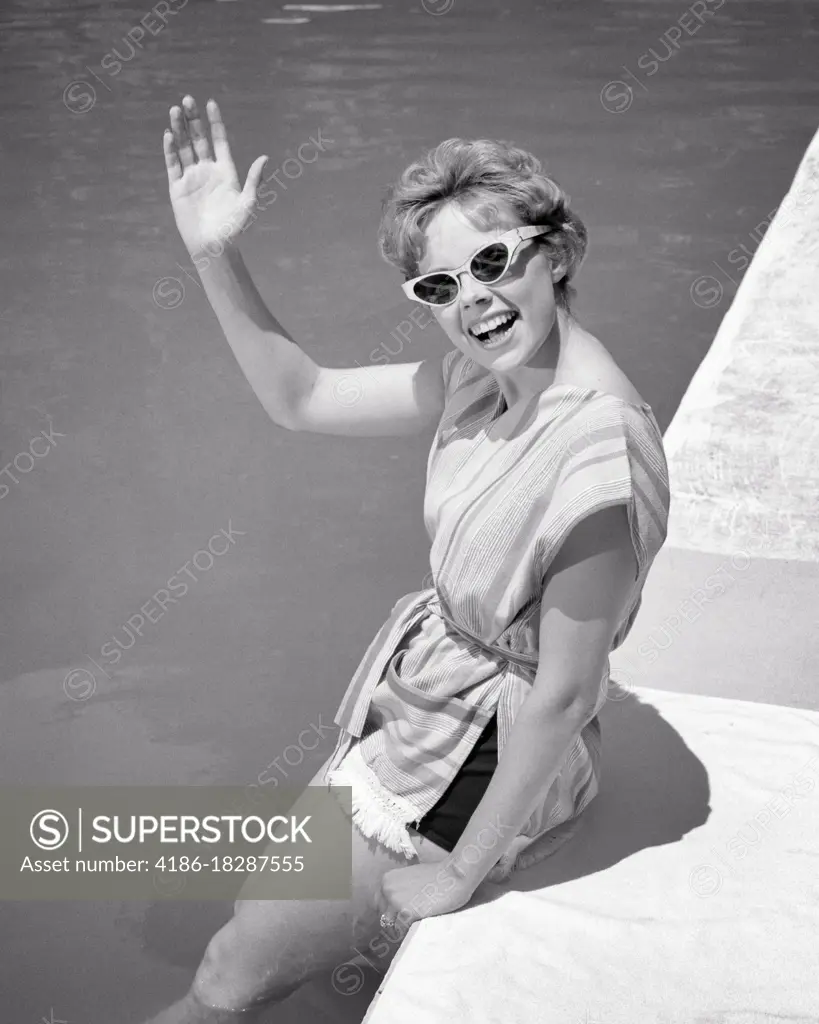 1960s smiling YOUNG woman sitting on edge of swimming pool waving greeting wearing sunglasses