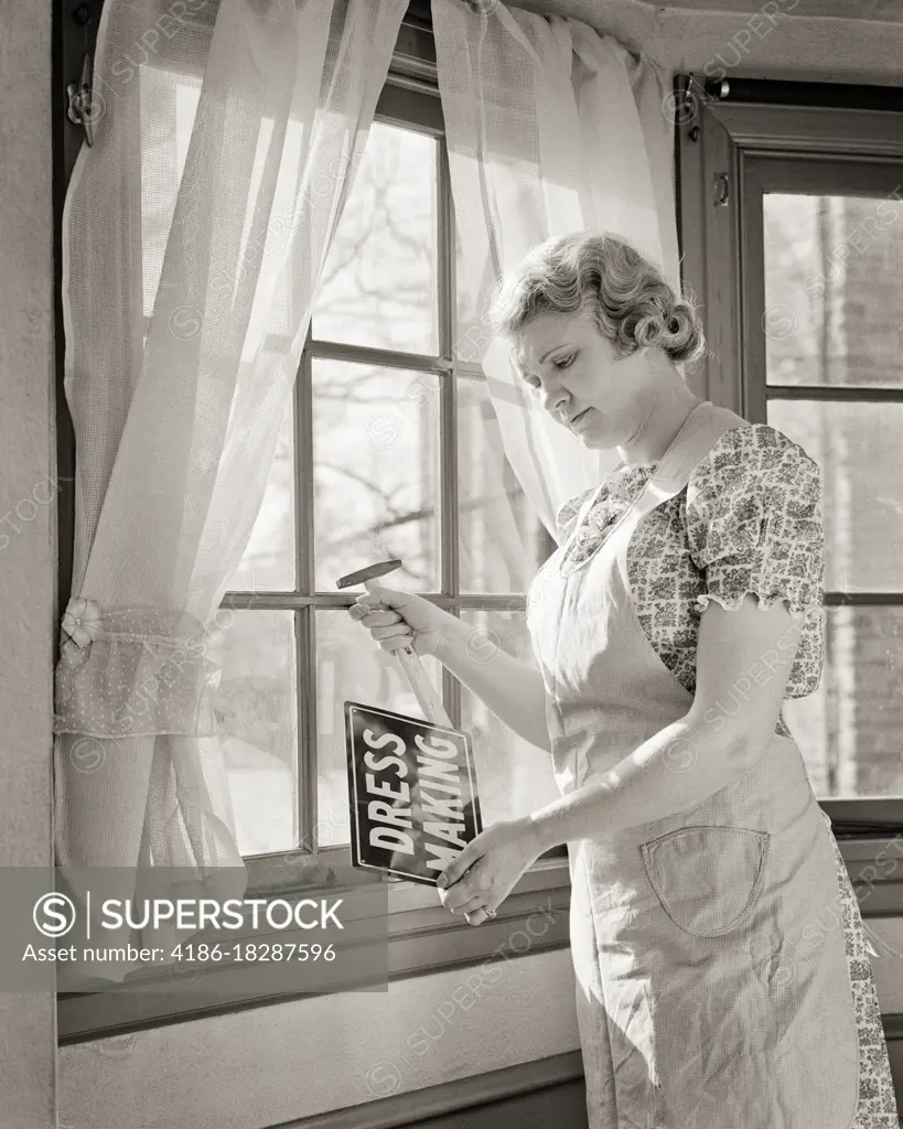 1940s MIDDLE AGED WOMAN PUTTING A DRESS MAKING SERVICES SIGN IN THE WINDOW OF HER HOME