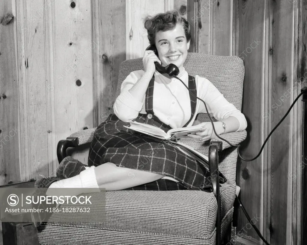 1950s TEEN GIRL SMILING TALKING ON THE TELEPHONE READING BOOK IN HER LAP WEARING BOBBY SOCKS LOAFERS SITTING IN EASY CHAIR