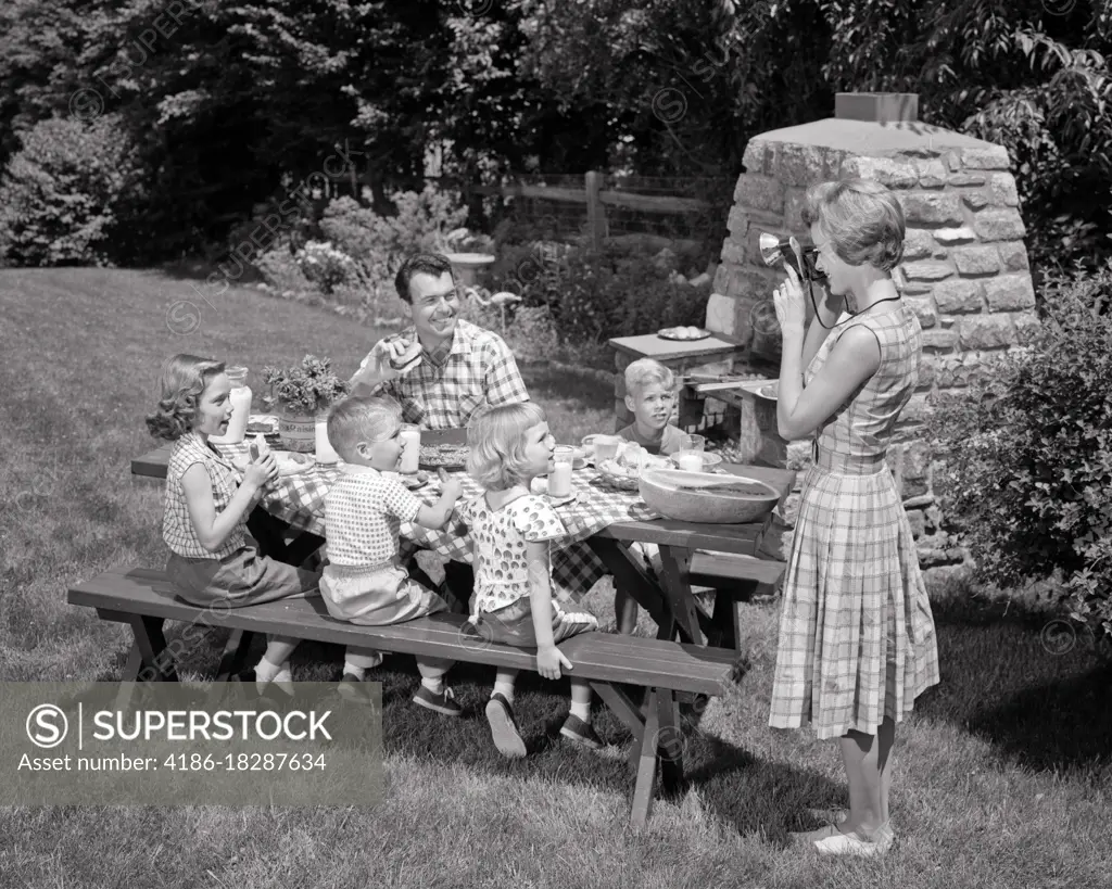 1960s MOTHER TAKING SNAPSHOT PHOTO OF FAMILY PICNIC BACKYARD BBQ KIDS AND DAD AT TABLE 