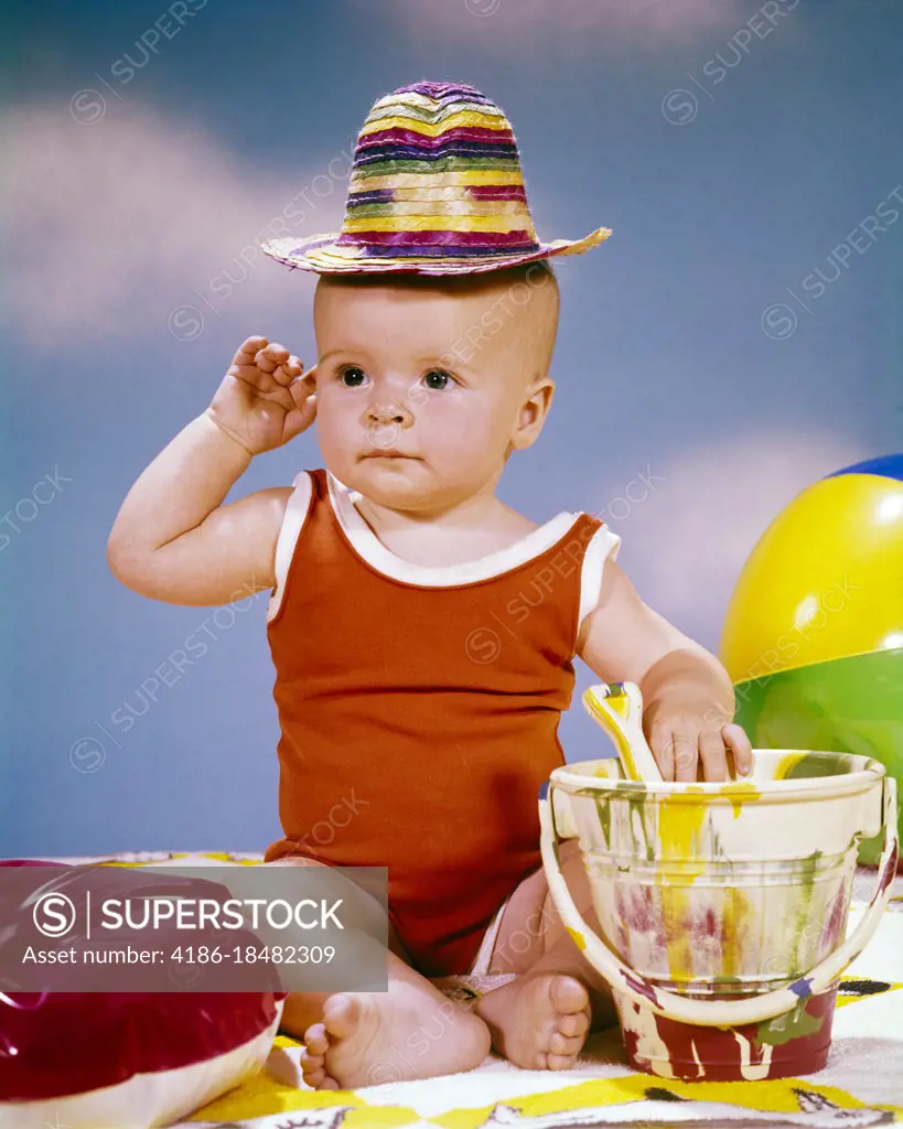 1950s 1960s BABY GIRL WEARING SILLY SMALL STRAW HAT RED BATHING SUIT WITH PLASTIC SAND PAIL ONE ARM RAISED SITTING ON BEACH SAND