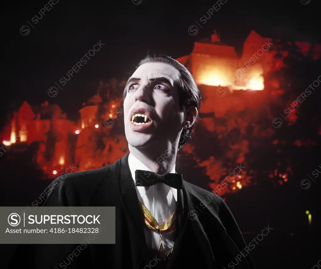 1970s MAN IN VAMPIRE MAKEUP AND COSTUME LOOKING UP MENACINGLY MOUTH OPEN SHOWING HIS BLOODTHIRSTY FANGS CASTLE BACKGROUND