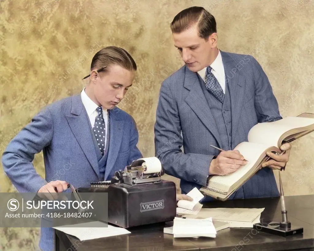 1920s BOOKKEEPER AND YOUNG ASSISTANT IN OFFICE USING LEDGER BOOK AND MANUAL ADDING MACHINE