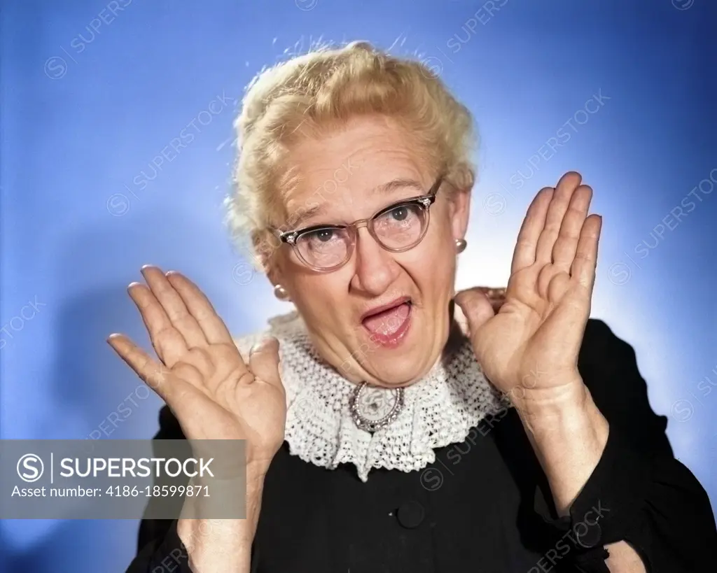 1960s ELDERLY WOMAN IN GRANNY GLASSES HOLDING HAND UP NEAR FACE WITH MOUTH  OPEN LOOKING AT CAMERA - SuperStock