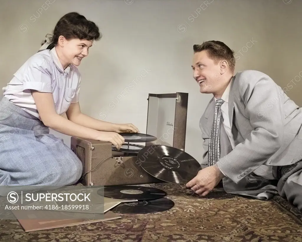 1950s TEENAGE SMILING COUPLE BOY AND GIRL SITTING ON FLOOR PLAYING VINYL MUSIC RECORDS ON PORTABLE PHONOGRAPH