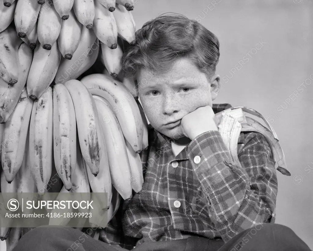 1930s 1940s DISTRESSED FRECKLE FACED BOY WITH STOMACH ACHE LOOKING AT CAMERA SITTING BY BUNCH OF BANANAS ONE HALF EATEN IN HAND