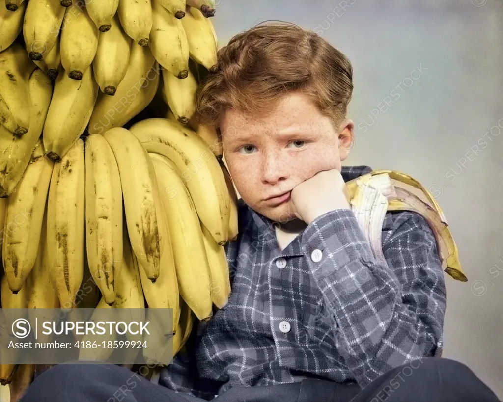 1930s 1940s DISTRESSED FRECKLE FACED BOY WITH STOMACH ACHE LOOKING AT CAMERA SITTING BY BUNCH OF BANANAS ONE HALF EATEN IN HAND