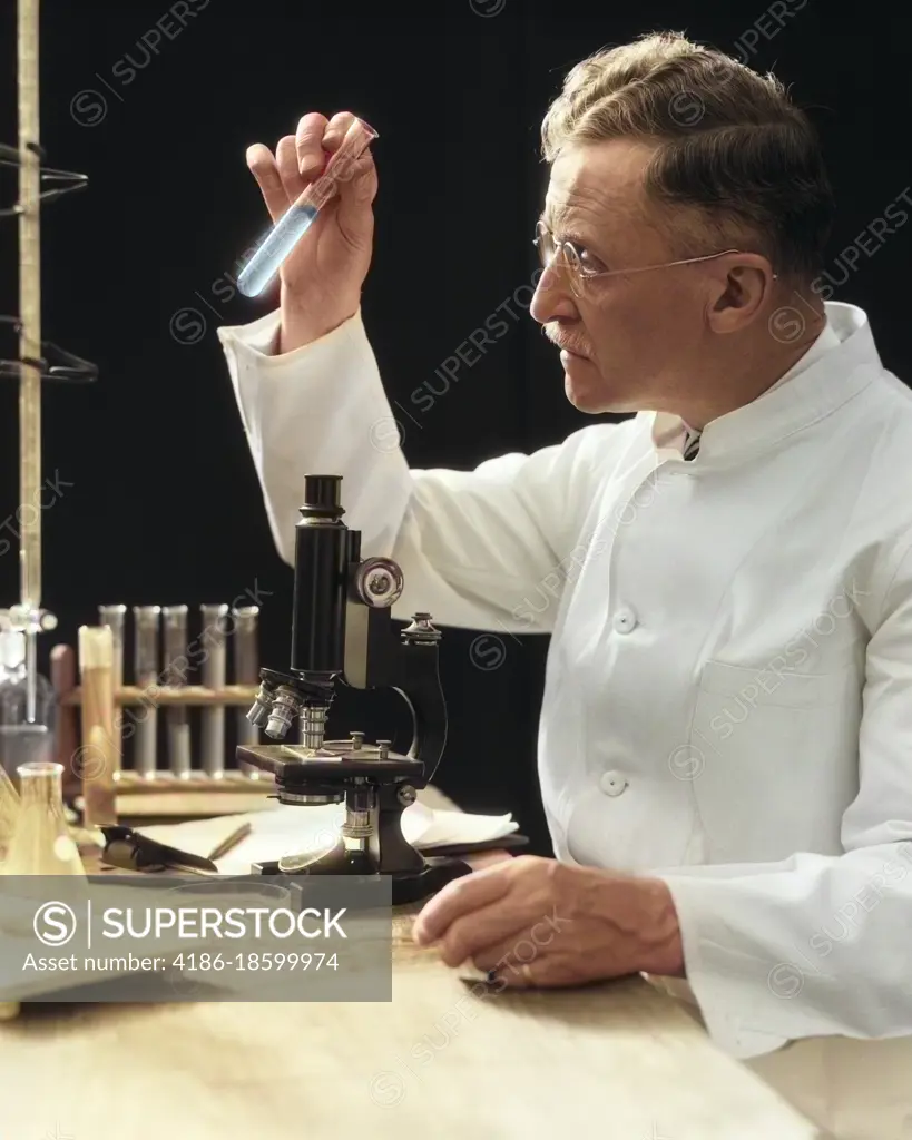 1920s 1930s 1940s MAN SCIENTIST LAB TECHNICIAN IN WHITE COAT LOOKING AT TEST-TUBE IN FRONT OF MICROSCOPE