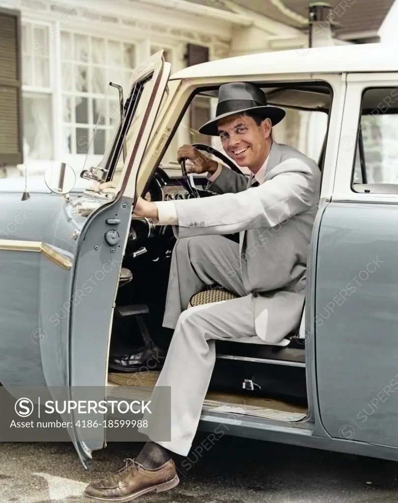 1950s SMILING MAN COMMUTER IN HAT SUIT TIE LOOKING AT CAMERA HOLDING OPEN DRIVER CAR DOOR STEPPING OUT RETURNING HOME AFTER WORK