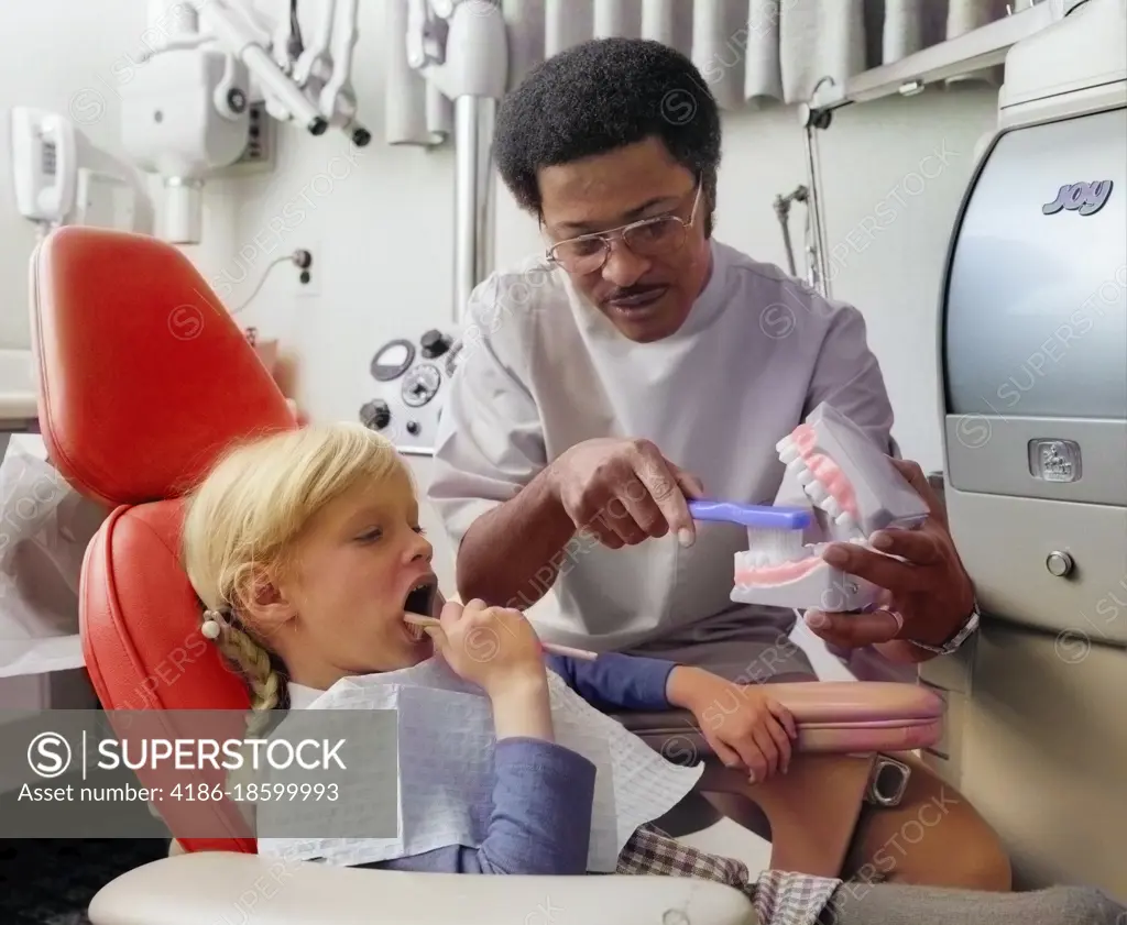 1970s AFRICAN AMERICAN MAN DENTIST TEACHING LITTLE BLOND CAUCASIAN GIRL HOW TO BRUSH HER TEETH USING LARGE MODEL OF JAW