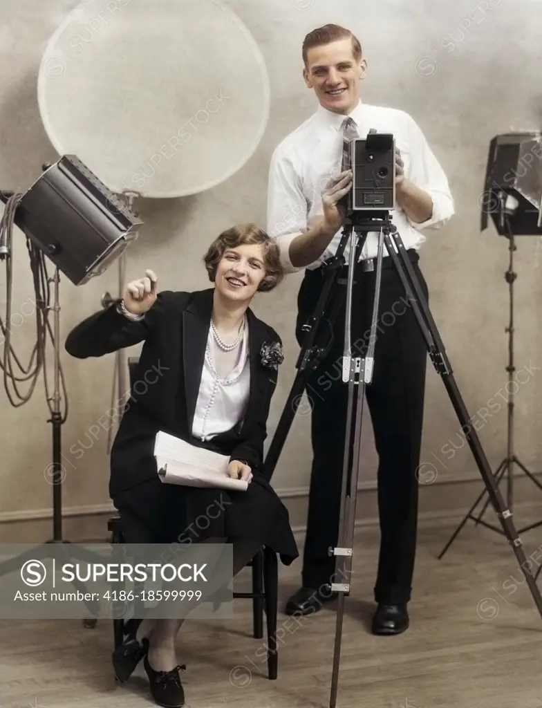 1920s PHOTOGRAPHER STANDING BEHIND MOVIE CAMERA IN STUDIO WITH ASSISTANT SITTING BESIDE HOLDING SCRIPT BOTH LOOKING AT CAMERA