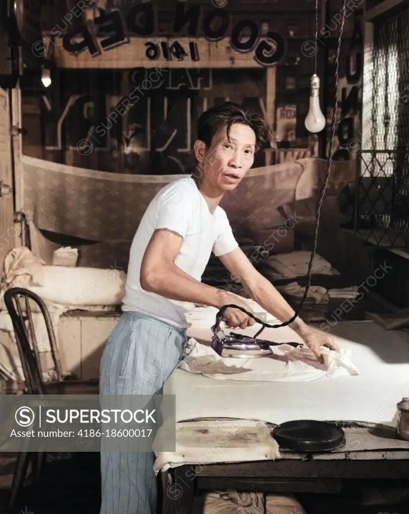 1920s CHINESE LAUNDRY MAN LOOKING AT CAMERA WORKING HARD IRONING CLOTHES BY HAND ON FLAT BOARD