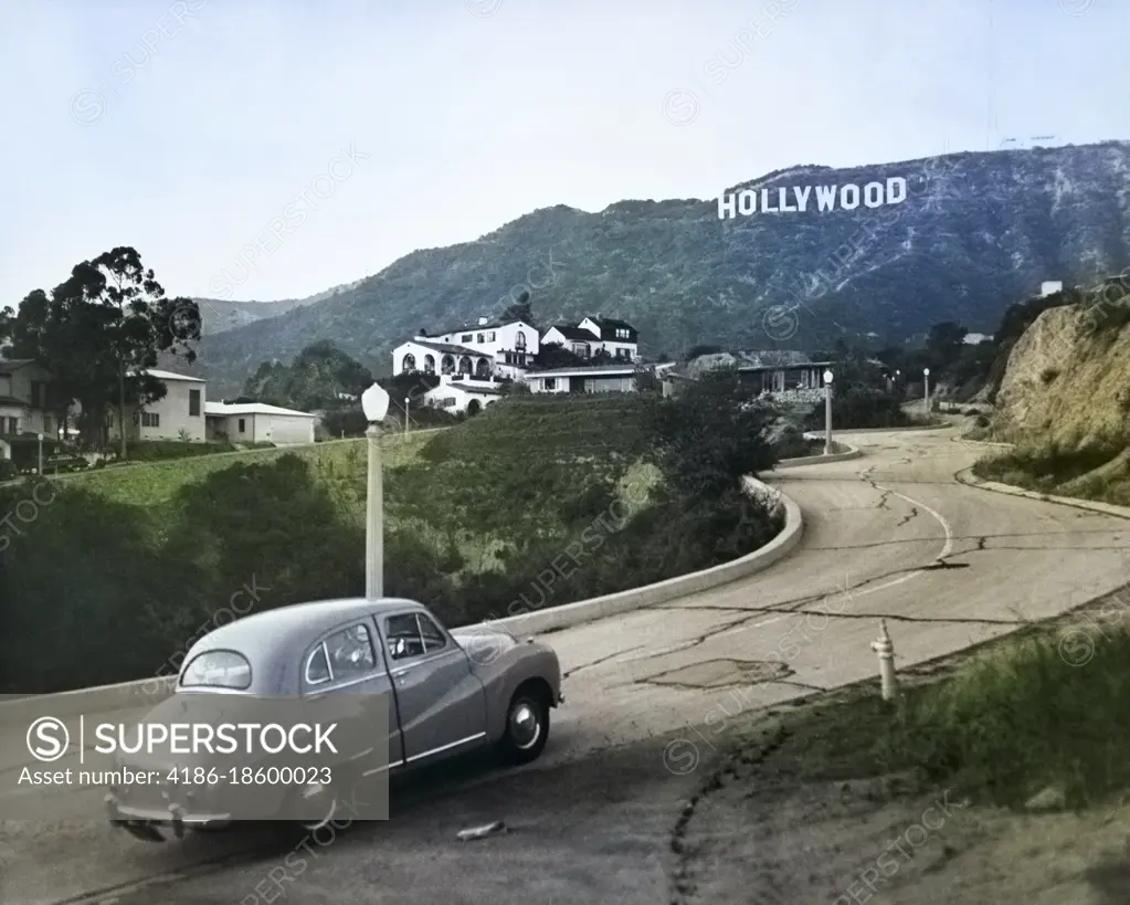 1950s AUSTIN CAR DRIVING UP ROAD IN THE HOLLYWOOD HILLS WITH HOLLYWOOD SIGN IN DISTANCE LOS ANGELES CALIFORNIA USA