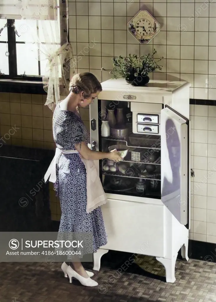 1920s 1930s BLOND WOMAN IN APRON SILK PARTY DRESS HIGH HEELS TAKING DISH FROM ELECTRIC KITCHEN REFRIGERATOR