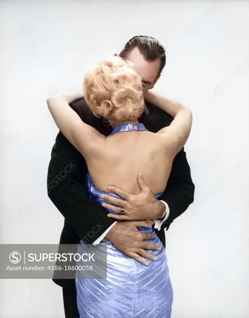 1950s 1960s KISSING COUPLE BLOND WOMAN BACKLESS SATIN EVENING DRESS MAN  EMBRACING FROM BEHIND - SuperStock