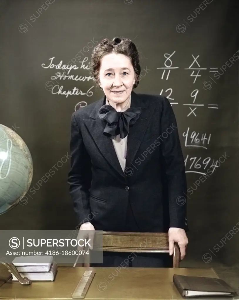 1940s SMILING SCHOOL TEACHER STANDING BEHIND HER DESK & IN FRONT OF THE BLACK BOARD WITH FRACTIONS