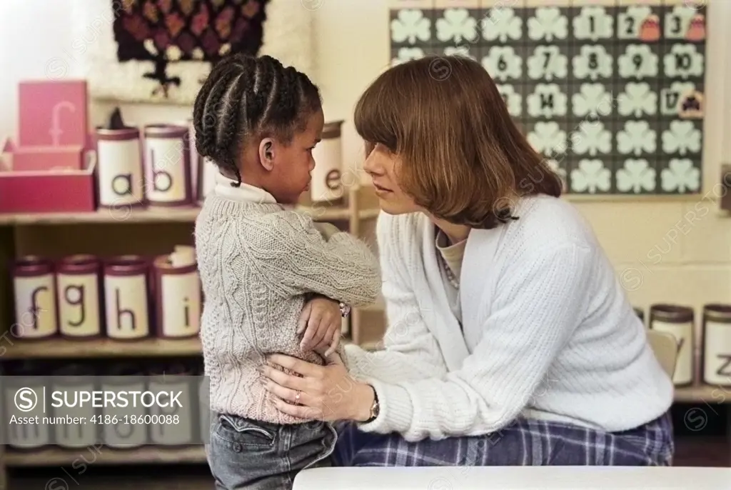 1980s WOMAN CAUCASIAN GRADE SCHOOL TEACHER BENDING DOWN TO COMFORT ANGRY UPSET AFRICAN AMERICAN LITTLE GIRL STUDENT