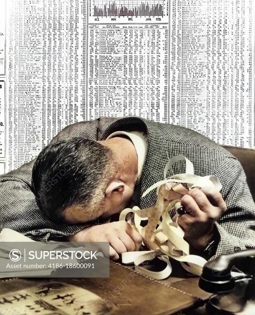 1940s MAN HEAD ON DESK HOLDING STOCK TICKER TAPE WITH NEWSPAPER STOCK PAGE BEHIND HIM