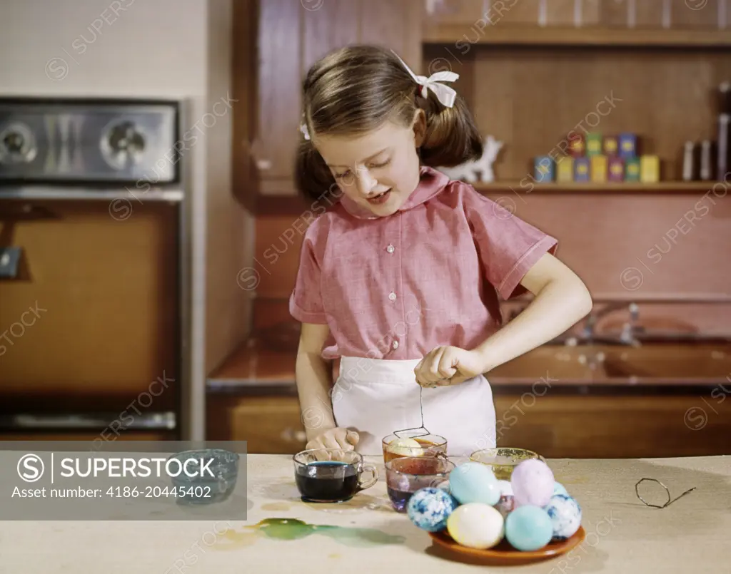 1960s SMILING GIRL WEARING PINK BLOUSE DYEING EASTER EGGS IN KITCHEN