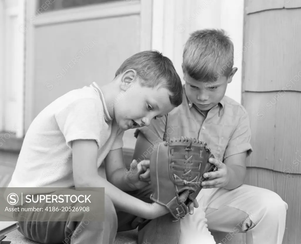 1970s TWO BOYS SERIOUSLY INSPECTING NEW LEATHER BASEBALL MITT SITTING ON PORCH