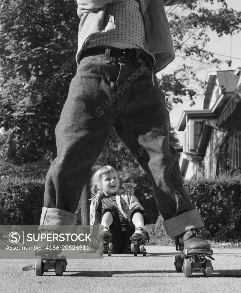 1950s GIRL FALLEN SITTING ON SIDEWALK WEARING METAL ROLLER SKATES SHOT THROUGH LEGS OF BOY ROLLED CUFF BLUE JEANS