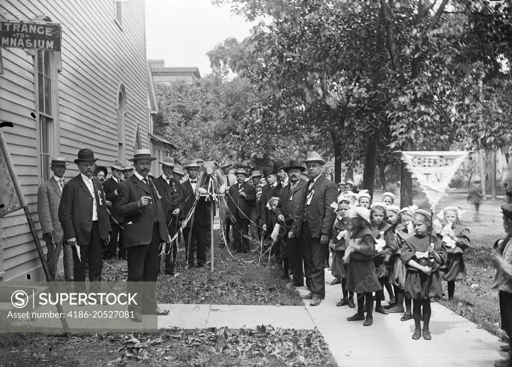 1890s TURN OF THE 20TH CENTURY GROUP OF SCHOOL CHILDREN WEARING UNIFORMS STANDING BY GROUP OF MEN HOLDING MAYPOLE RIBBONS