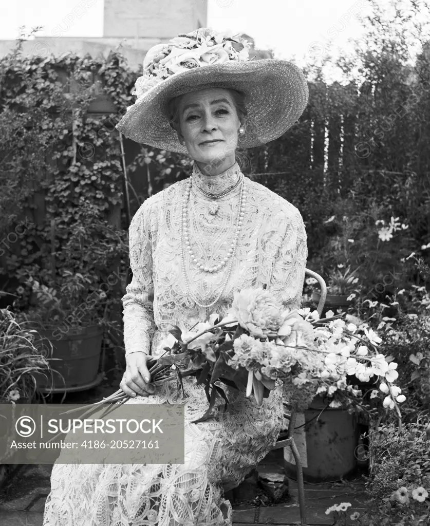 1970s SENIOR WOMAN SITTING IN GARDEN HOLDING BOUQUET FLOWERS WEARING ANTIQUE LACE DRESS BIG HAT SMILING LOOKING AT CAMERA