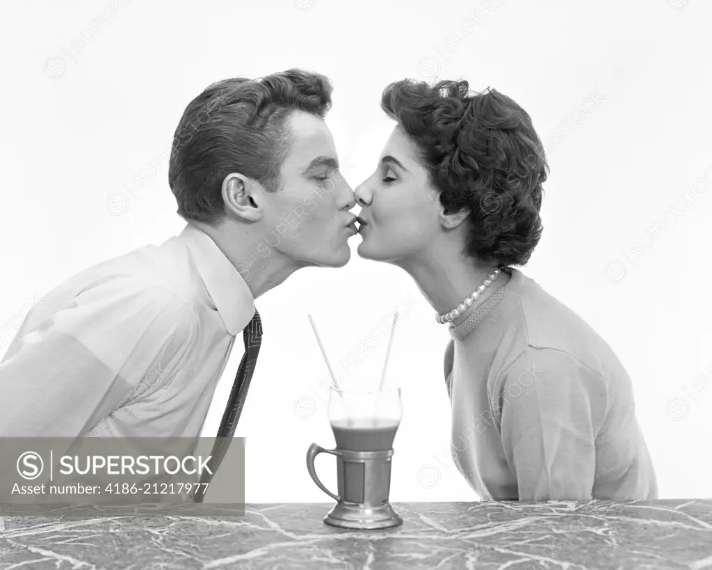 1950s COUPLE MAN WOMAN KISSING PROFILE ROMANCE DATE AT MARBLE COUNTER ONE DRINK MALT SHAKE WITH TWO STRAWS SHARE TEENS TEENAGERS