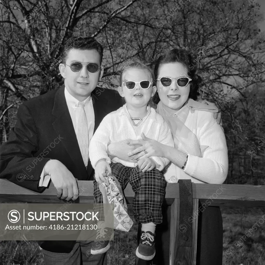 1950s FAMILY FATHER MOTHER DAUGHTER AT FENCE WEARING DARK SUNGLASSES LOOKING AT CAMERA