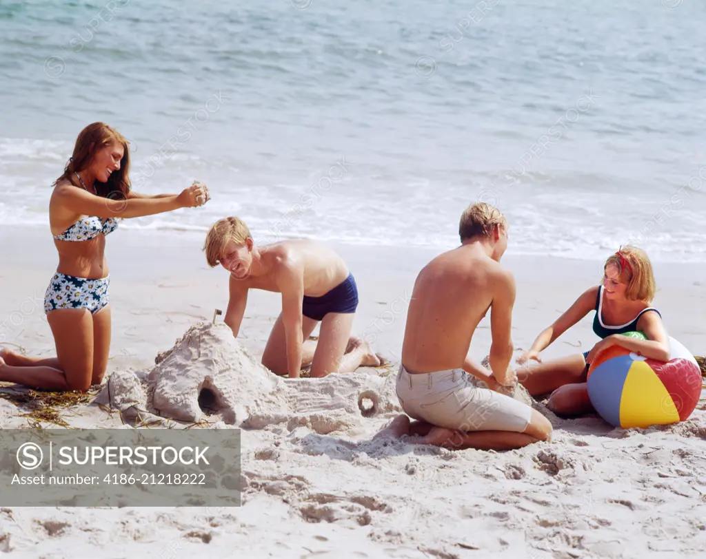 1960s 1970s TWO TEENAGE COUPLES ON BEACH BUILDING SANDCASTLE AND PLAYING WITH BALL