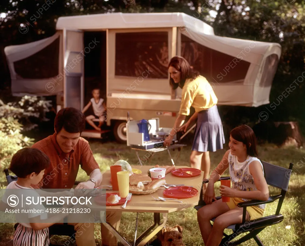 1970s FAMILY CAMPING VACATION CAMPSITE MOTHER COOKING ON CAMP STOVE FATHER AND KIDS AT TABLE EATING