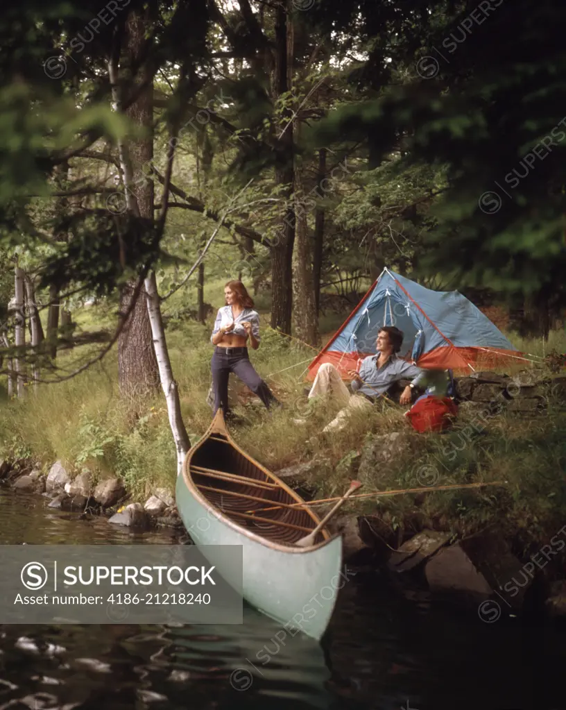 1970s COUPLE AT A FOREST CAMP CAMPSITE WITH TENT AND CANOE IN STREAM 